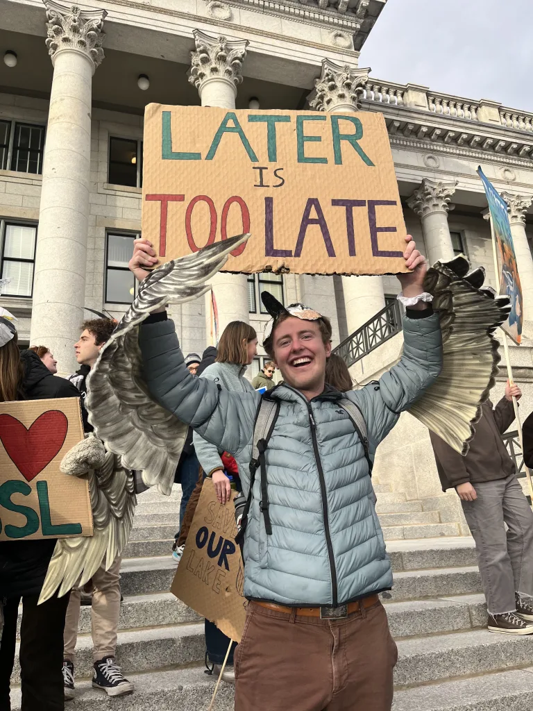 A smiling protester stands on the steps of a grand, columned building, holding a handmade sign that reads “LATER IS TOO LATE” in bold, colorful letters. They wear a light blue puffer jacket and brown pants, with digitally added bird wings extending from their arms—symbolizing urgency, transformation, or freedom. Around them, other demonstrators hold signs with messages about climate action, including one partially reading “OUR PLANET IS…” and another with a heart and the letters “SL.” The scene captures a spirited, creative moment in a climate protest.
