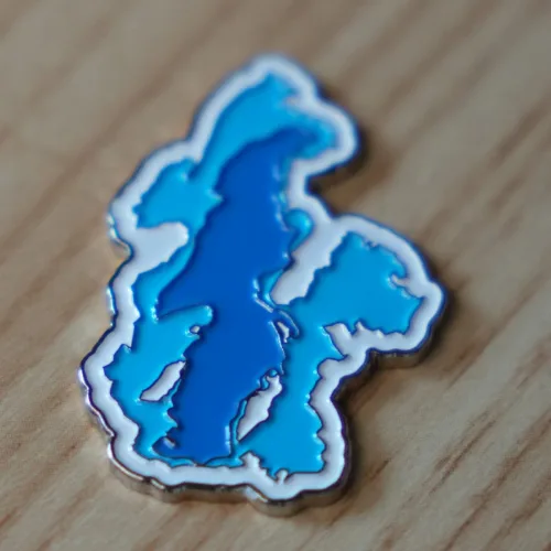 A stylized enamel pin shaped like the Great Salt Lake, rendered in layered blues that evoke depth and fragility. Its contours trace the lake’s shrinking edges, set against a warm wooden surface—an emblem of place, memory, and ecological urgency.